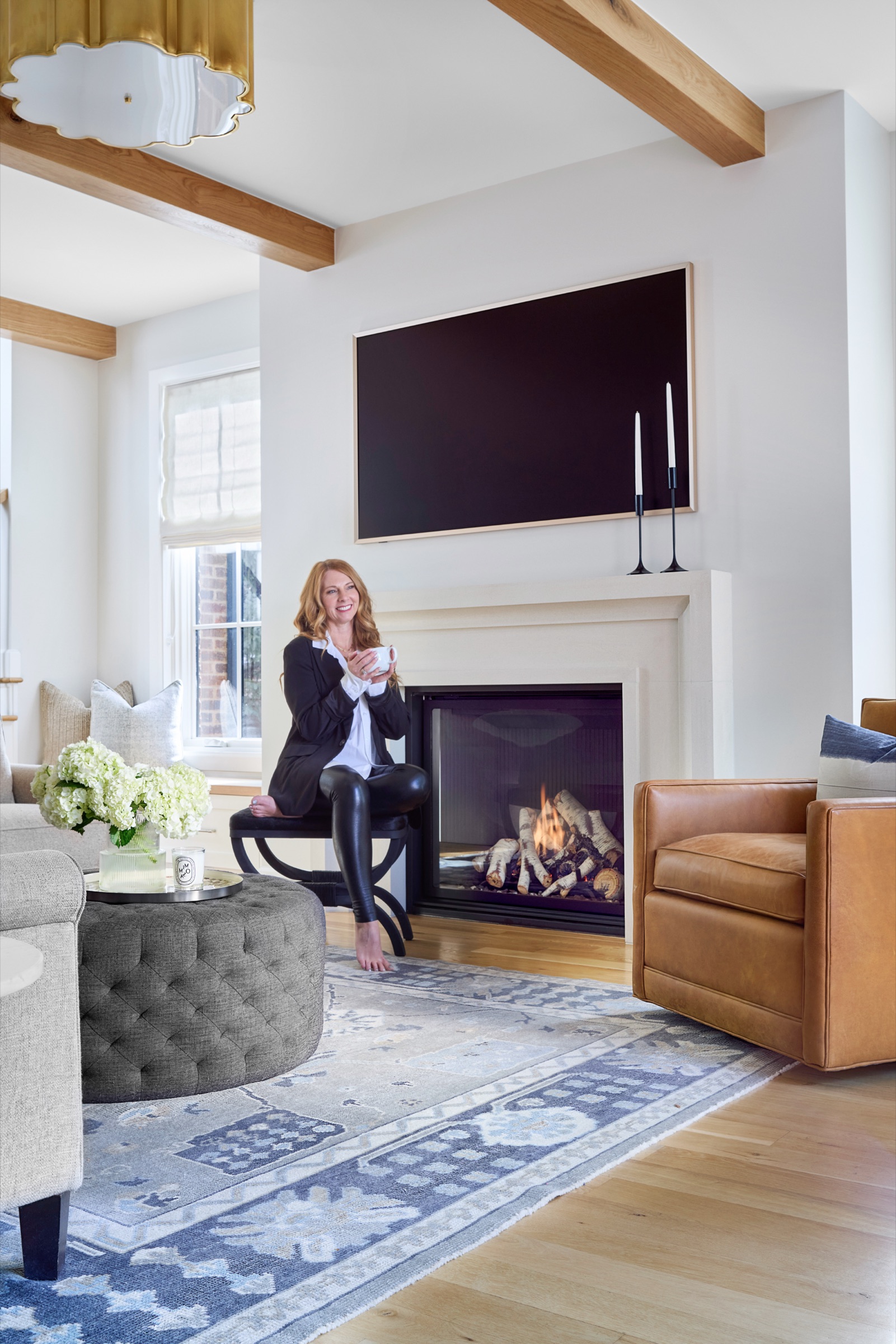 photo of owner and principal interior designer, Kendra Stewart, owner at E+E Design consulting holding a mug looks like you could have a conversation with her. She is sitting next to a lit fireplace with white cast surround, black frame tv, camel leather chair, blue printed rug and gray tufted ottoman with beautiful white hydrangea flowers, her favorite. 