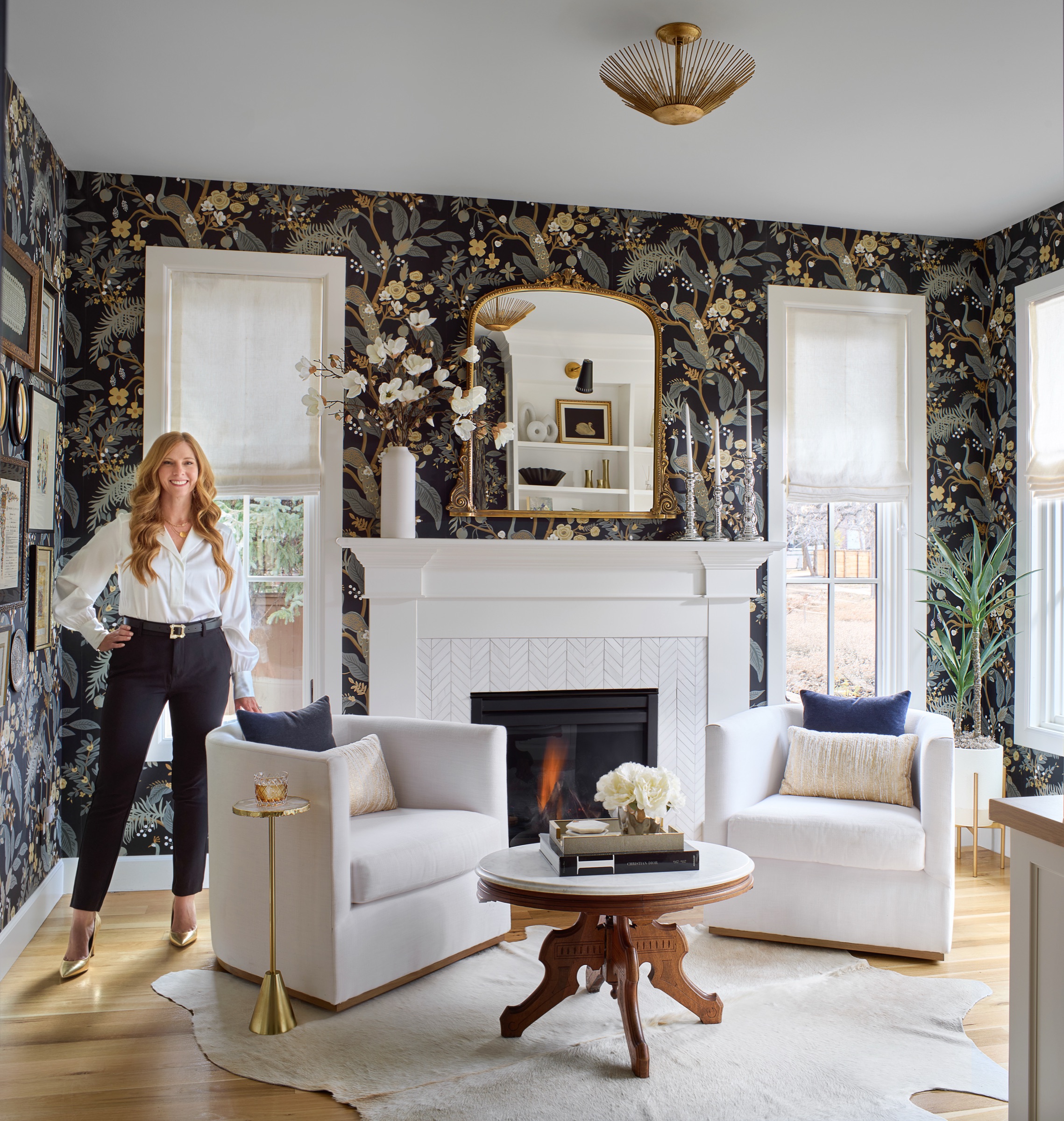 Kendra Stewart, owner and principal interior designer at E+E Design Consulting, in a white swivel chair in a sitting room office with peacock wallpaper, marble chevron fireplace, and gold mirror