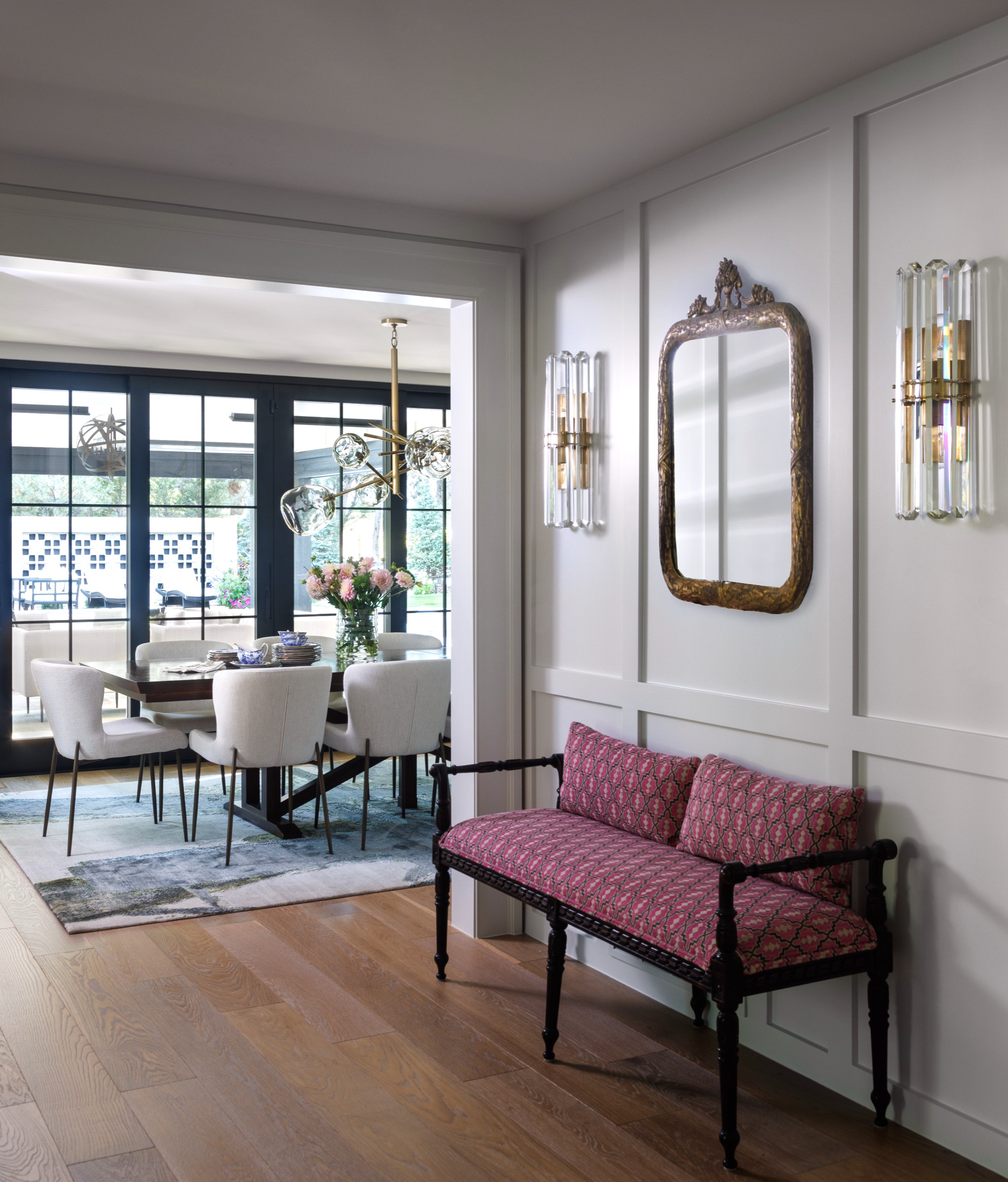 photo of hallways with white paneling, an antique bench with pink printed cushions, mirror above with crystal sconces and in the background its showing a dining table with organic rug, white dining chairs and gold and glass chandelier and a view to the pool and patio outside through the sliding glass doors
