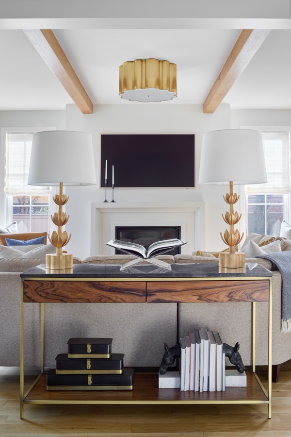 beautiful photo of a sofa table in a living room with the fireplace in the background. the sofa table has two gold lamps with white lampshades and an acrylic book stand with a book displayed. the sofa table has black marble top, walnut drawers and a gold frame with a shelf at the bottom holding black and gold boxes and books with black horse bookends
