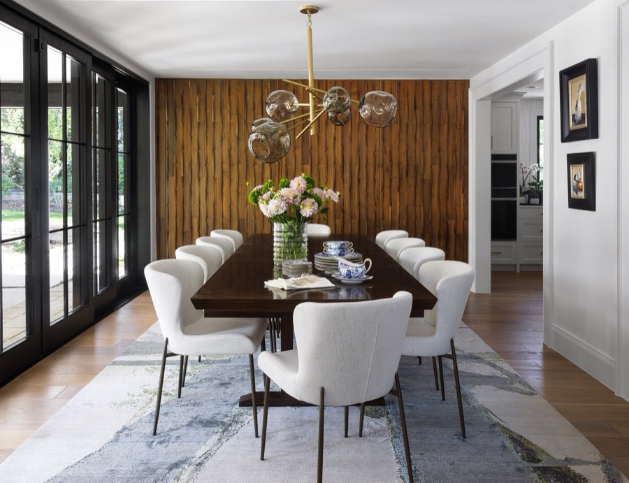 photo of a dining room table with white chairs, organic rug with blues and greens, and a gold and glass ball chandelier. on the left are sliding black glass doors to the outside patio and pool area, in the background are walnut 3D wall paneling with hidden doors to the storage china cabinet and pool guest bathroom. on the right is the walkway to the kitchen and you can see the Wolf modern double ovens.