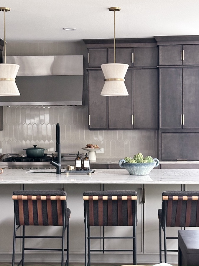 Kitchen island with dark cabinetry, marble-look countertop, black faucet, pendant lights, leather bar stools, and textured backsplash tile.