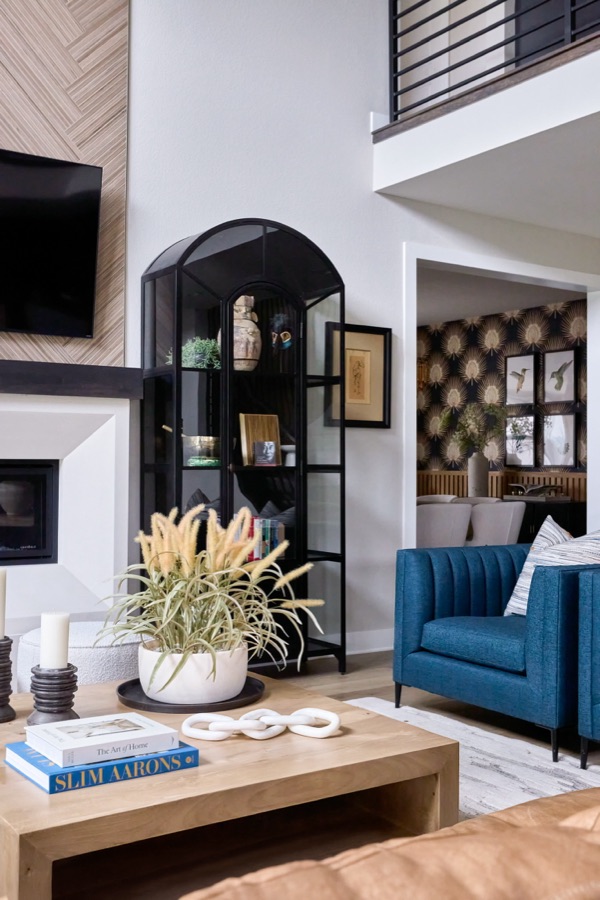 Living room detail with blue chair, arched black display cabinet, fireplace, coffee table, and dining room with wallpaper in the background.