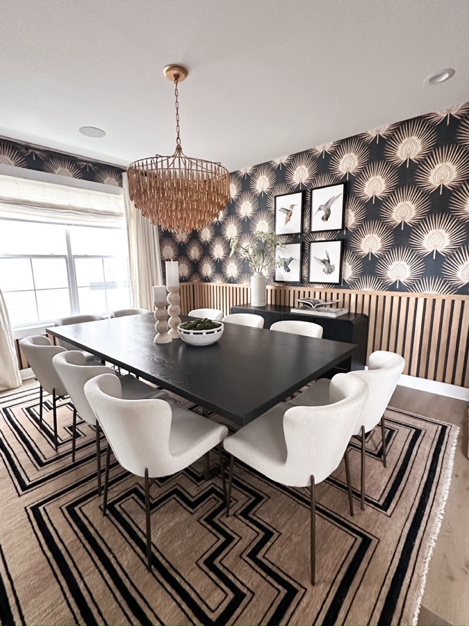 Dining room with black and cream wallpaper, wood slat wall paneling, black dining table, white chairs, geometric rug, and chandelier.
