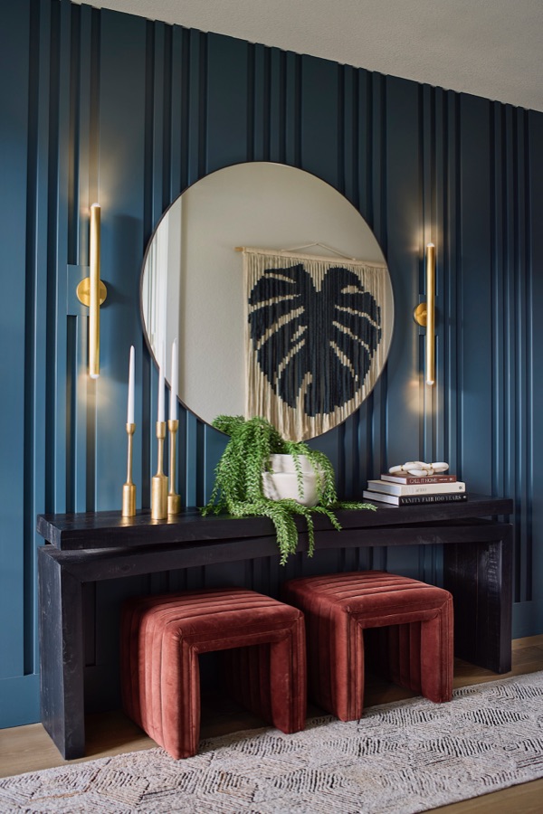 Entryway with teal paneled wall, round mirror, brass sconces, black console, rust velvet stools, and woven wall art.