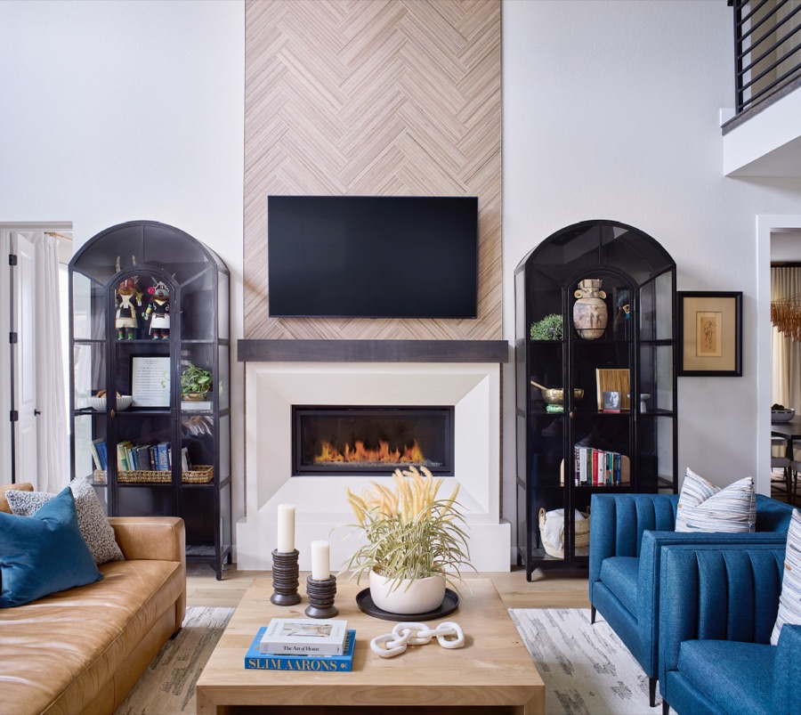 Living room fireplace wall with cast-stone surround, herringbone wall detail, arched black display cabinets, blue chairs, and tan sofa.