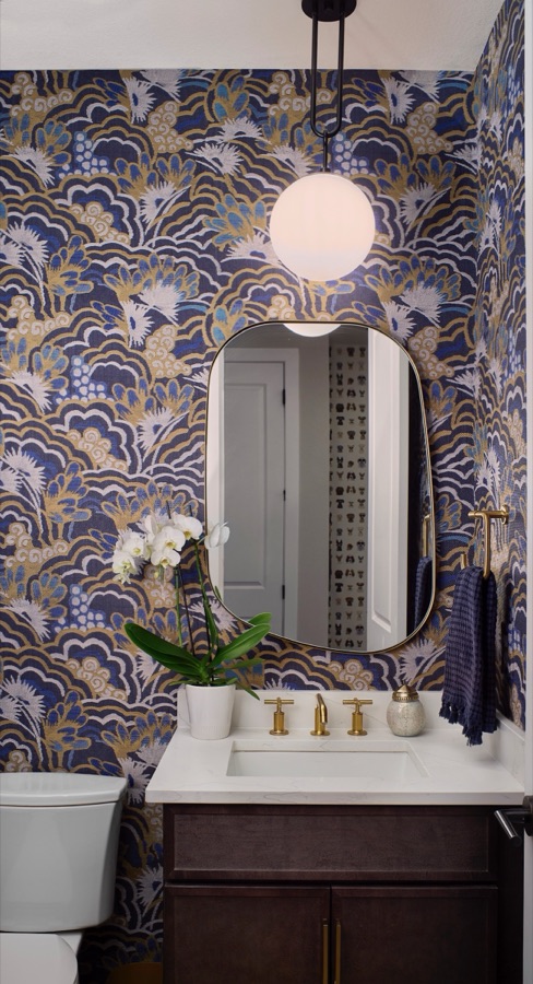 Powder bathroom with blue and gold patterned wallpaper, oval mirror, brass faucet, pendant light, and dark vanity.