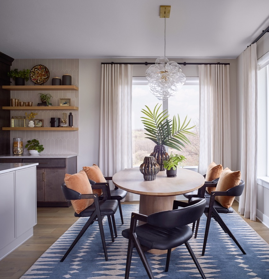 Breakfast nook with round wood table, black chairs, orange pillows, blue rug, open shelves, and large window.