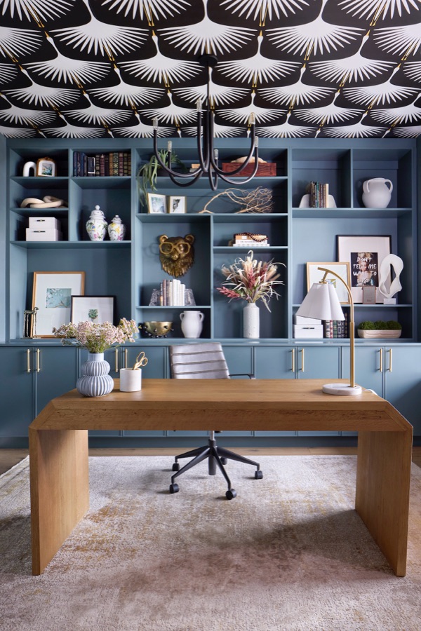 Home office with blue custom built-ins, wood desk, black and white wallpapered ceiling, and brass-accent desk lamp.