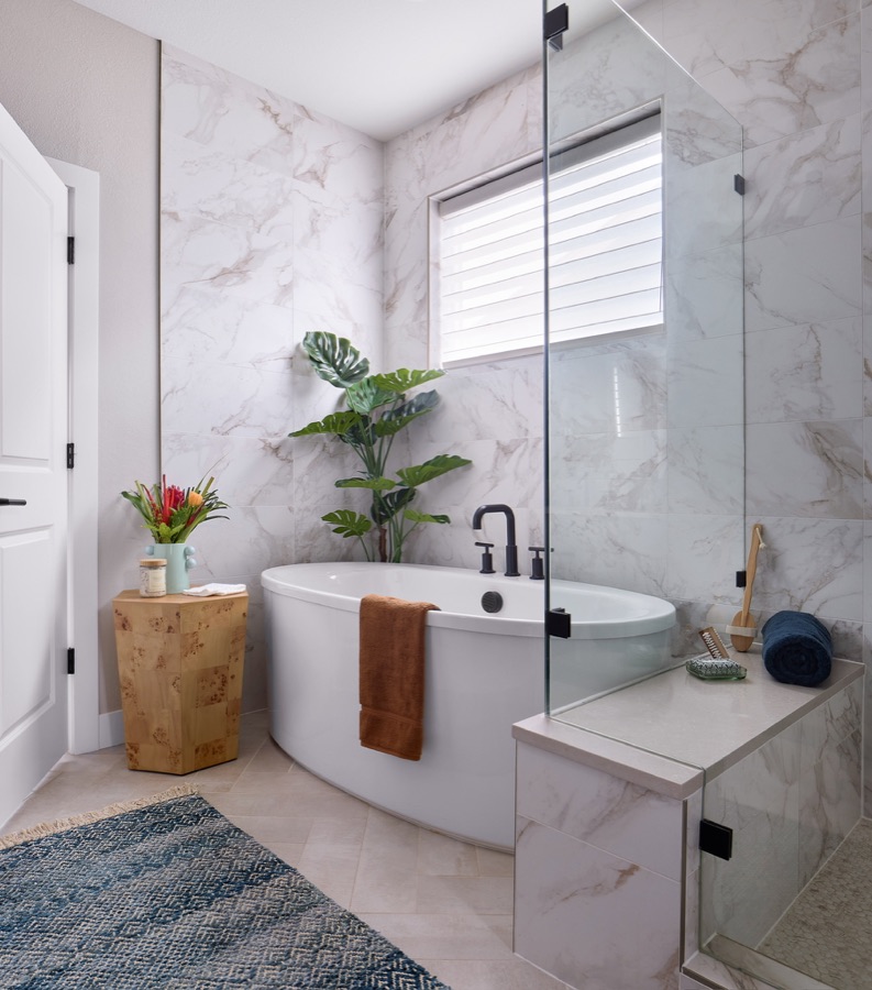 Primary bathroom with freestanding tub, glass shower, marble-look wall tile, black fixtures, window, and blue rug.